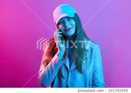 Woman talking on cellphone while standing against pink neon wall and looking smiling directly at camera, wearing leather jacket and baseball cap. Woman talking on cellphone while standing against pink neon wall and looking smiling directly at camera, wearing leather jacket and baseball cap. 101742840