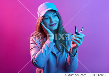 Cute attractive woman wearing stylish outfit and baseball cap holding pomades with two different shades, posing isolated over pink neon background. 101742852