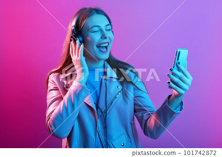 Emotional beautiful energetic young female with headphones listening to music, holding smartphone in one hand, recording video, opening mouth widely, singing with pleasure. Technology cocnept. 101742872