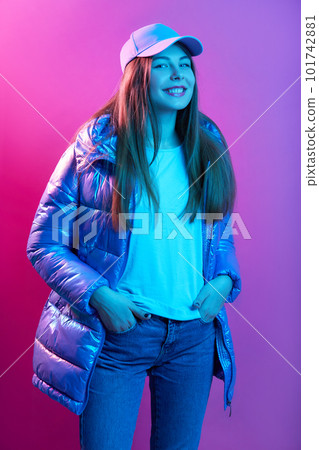 Close up portrait of beautiful woman in fashion pose looking aside in winter clothing, lady wearing jeans, cap and puffer jacket, smiling girl with hands in pocket, girl looks happy and satisfied. 101742881