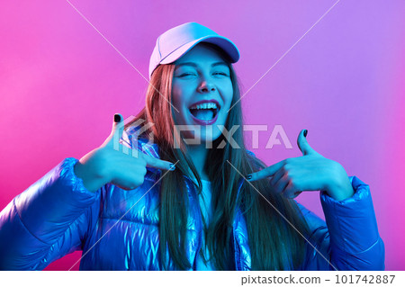 Confident happy excited female wearing jacket and baseball cap, standing against pink neon wall, yelling something and pointing to herself with index fingers, keeps mouth widely opened. 101742887