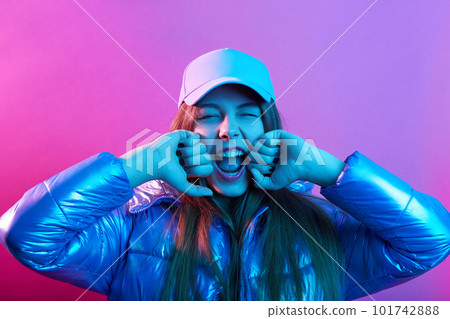 Young attractive girl in baseball cap and jacket, yelling happily, keeping fists on cheeks, expressing positive and gladness, posing isolated over pink neon background. Young attractive girl in baseball cap and jacket, yelling happily, keeping fists on cheeks, expressing positive and gladness, posing isolated over pink neon background. 101742888