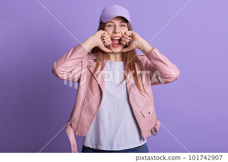 Horizontal shot of woman with long fair hair poses in studio dressed white shirt, pink leather jacket and cap, attractive young female screaming something, keeps fists near her face against lilac wall 101742907