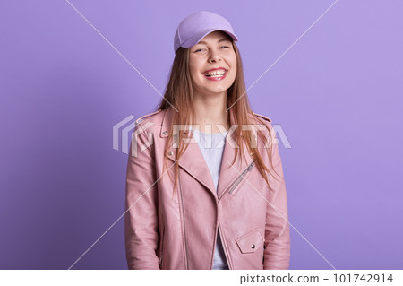 Horizontal shot of fashion woman with long fair hair posing in studio against lilac wall, lady dressedrosy leather jacket and cap, female standing with toothy smile and happy facial expression. 101742914