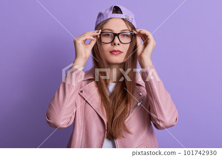 Close up portrait of concentrated serious young female touching eyeglasses frame with fingers, trying new eyewear, looking directly at camera, wearing stylish clothes. Youth and trend concept. 101742930