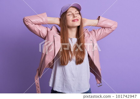 Horizontal shot of adorable relaxed cute young female closing eyes, putting hands behind head, enjoying free time, being fond of spring, wearing t shirt, leather jacket and cap. Youth concept. Horizontal shot of adorable relaxed cute young female closing eyes, putting hands behind head, enjoying free time, being fond of spring, wearing t shirt, leather jacket and cap. Youth concept. 101742947