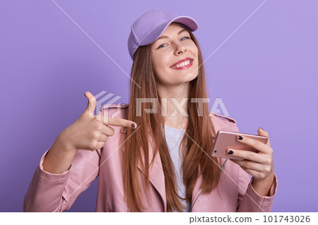 Indoor shot of positive delighted charming female with long hair, wearing cap, pink leather jacket and t shirt, holding smartphone, showing direction with forefingers. Youth and free time concept. 101743026