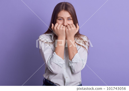 Horizontal indoor shot of attractive beautiful young female covering her mouth with both fists, standing isolated over lilac background in studio, having cunning facial expression. Youth concept. Horizontal indoor shot of attractive beautiful young female covering her mouth with both fists, standing isolated over lilac background in studio, having cunning facial expression. Youth concept. 101743056