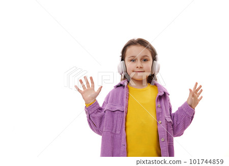 Caucasian happy adorable little girl in bright yellow sweater and purple jacket, wearing wireless headphones, looks and camera and shows her hands palms up, isolated over white background. Copy space 101744859