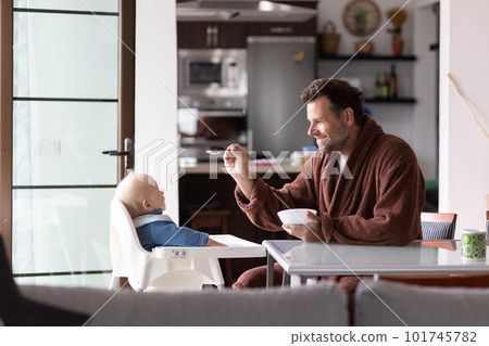 Father wearing bathrope spoon feeding hir infant baby boy child sitting in high chair at the dining table in kitchen at home in the morning 101745782