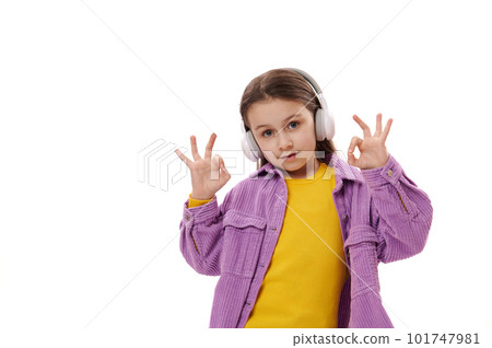 Elementary age child, lovely little school girl in casual clothes, enjoys listening music on wireless headphones, gestures with her hands, shows OK sign, smiles at camera, isolated white background Elementary age child, lovely little school girl in casual clothes, enjoys listening music on wireless headphones, gestures with her hands, shows OK sign, smiles at camera, isolated white background 101747981