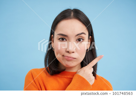 Close up portrait of beautiful modern girl, smirks skeptical, points right, shows banner, stands over blue background 101748563
