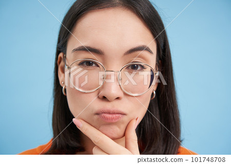 Close up portrait of asian girl in glasses, squints with suspicious face, thinking, stands over blue background 101748708