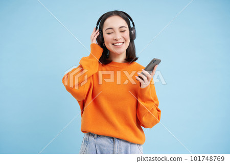 Beautiful young woman in headphones with mobile phone, listens to music in earphones and smiles, blue background 101748769