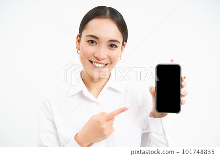 Portrait of businesswoman showing smartphone screen, pointing at mobile phone and smiling, recommending application, standing over white background 101748835