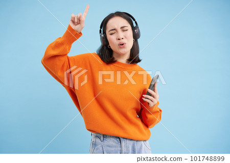 Beautiful korean woman in headphones, dancing, listening music on mobile phone app, smiling, posing over blue background Beautiful korean woman in headphones, dancing, listening music on mobile phone app, smiling, posing over blue background 101748899