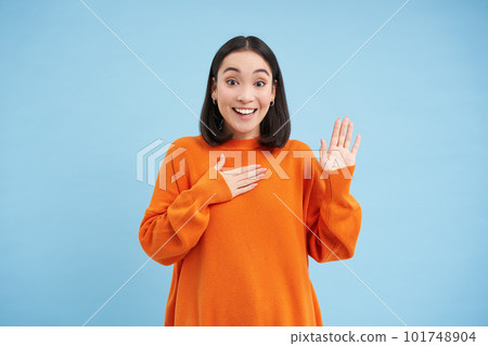 Smiling asian woman press arm to heart and raises one hand, introduces herself, stands over blue background Smiling asian woman press arm to heart and raises one hand, introduces herself, stands over blue background 101748904