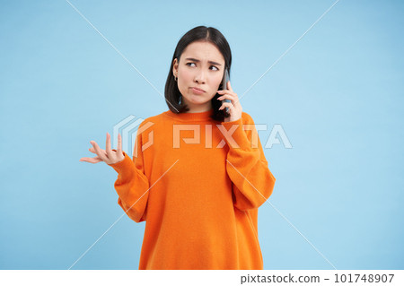 Sad and confused asia girl shrugs, talks on cellphone, answers telephone call with puzzled face, stands over blue background Sad and confused asia girl shrugs, talks on cellphone, answers telephone call with puzzled face, stands over blue background 101748907