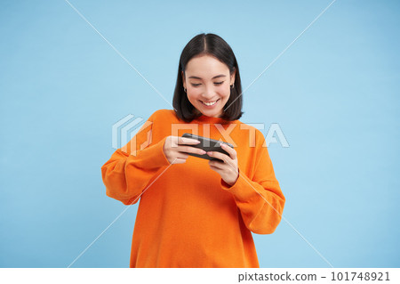 Happy smiling girl, 25 years, plays mobile video games, holds smartphone in both hands, stands against blue background 101748921