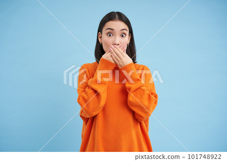 Portrait of shocked korean woman gasps, closes her mouth with hands and look speechless at camera, stands over blue background 101748922