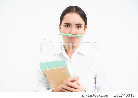 Funny korean woman with notebooks, holds pen with lips and nose and makes faces, stands over white studio background Funny korean woman with notebooks, holds pen with lips and nose and makes faces, stands over white studio background 101749013