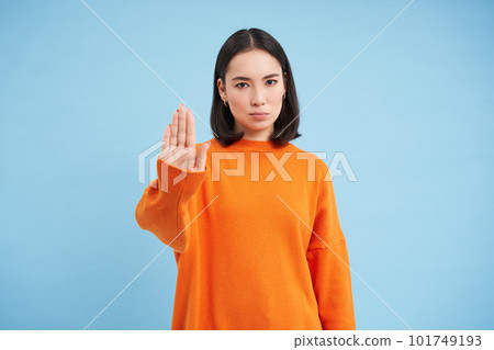 Stop. Serious and confident korean female model, extends hand, shows taboo, rejection gesture, refuse smth, stands over blue background 101749193