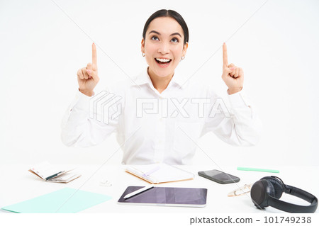 Business and profession. Portrait of asian woman in workplace, sits in office and points up, shows banner advertisement, white background Business and profession. Portrait of asian woman in workplace, sits in office and points up, shows banner advertisement, white background 101749238