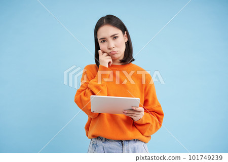 Complicated asian girl, stands with digital tablet with frowning, puzzled face, poses over blue background Complicated asian girl, stands with digital tablet with frowning, puzzled face, poses over blue background 101749239