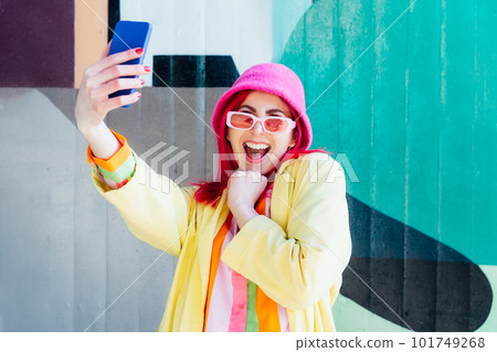Excited pink hair woman screaming while taking a selfie photo outdoors. Emotional hipster fashion woman in bright clothes, pearl pink sunglasses, bucket hat taking selfie photo on the phone camera Excited pink hair woman screaming while taking a selfie photo outdoors. Emotional hipster fashion woman in bright clothes, pearl pink sunglasses, bucket hat taking selfie photo on the phone camera 101749268