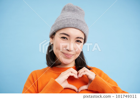 Close up of lovely asian girl in beanie, shows heart sign, looks cute, stands over blue studio background 101749311