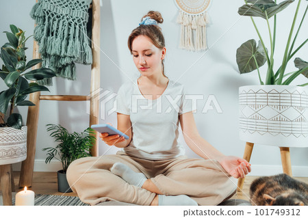 Young healthy woman using phone for setting mobile app for meditation at home, sitting in lotus position on floor cushion with relaxed cat pet in modern Scandi interior home. Meditating, relaxed 101749312