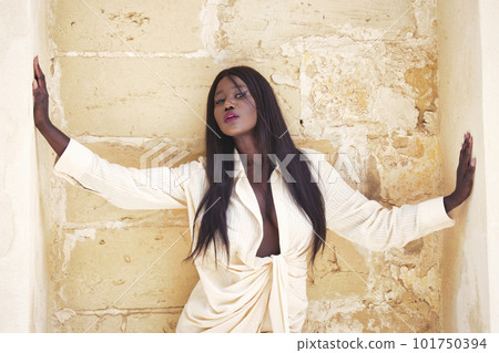 A beautiful black African woman posing in front of a weathered, rustic wall outside looking at the camera with a coy look on her face - upper body in frame 101750394