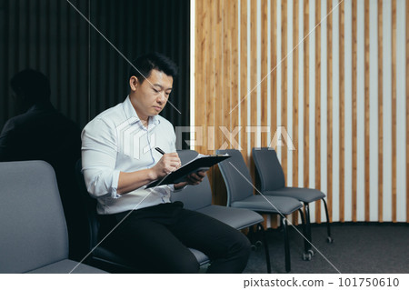 A young Asian man sits in the lobby and fills out a survey questionnaire in the medical center, in the office for work, interview, arrival at the hotel. A young Asian man sits in the lobby and fills out a survey questionnaire in the medical center, in the office for work, interview, arrival at the hotel. 101750610