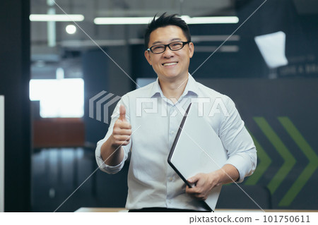 Portrait of a successful young Asian male programmer standing in a modern office with a notebook in his hands, smilingly looking at the camera, showing the super finger. Portrait of a successful young Asian male programmer standing in a modern office with a notebook in his hands, smilingly looking at the camera, showing the super finger. 101750611