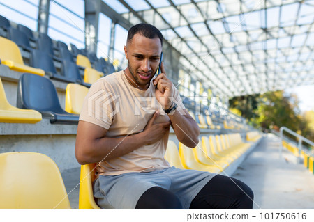 A young African American male athlete sits in the stadium, holds his hand to his chest and calls the doctor, the ambulance, the hospital for help. Suffers from pain, heart attack, stroke. A young African American male athlete sits in the stadium, holds his hand to his chest and calls the doctor, the ambulance, the hospital for help. Suffers from pain, heart attack, stroke. 101750616