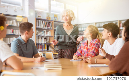 Adult female teacher helping pupils preparing for classes in library 101757078