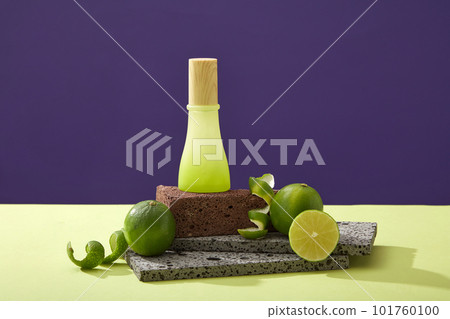 Glass bottle unlabeled on stone podium, halves of lime and spiral peel on color background. Mockup scene for cosmetic of lime extract - rich in vitamin C and nutrients for effective skin care 101760100
