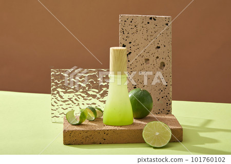 Front view of green glass bottle with wooden cap on brick podium, halves of lime and spiral lime peel on brown background. Mockup scene for product with lime ingredient. Space for design 101760102