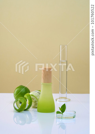 Front view of green bottle with wooden cap unlabeled, lab glassware, fresh limes and spiral peel decorated on color background. Mockup for product with ingredients from lemon essential oil. 101760522