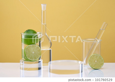 Transparent podium for display cosmetic product, lab glassware containing colorless liquid and fresh limes decorated on yellow background. Laboratory equipment and research natural extracts from lime 101760529