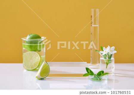 Transparent podium, fresh limes, flower and leaves decorated in lab glassware on yellow background. Blank space for display cosmetic with ingredient from lime. Front view 101760531