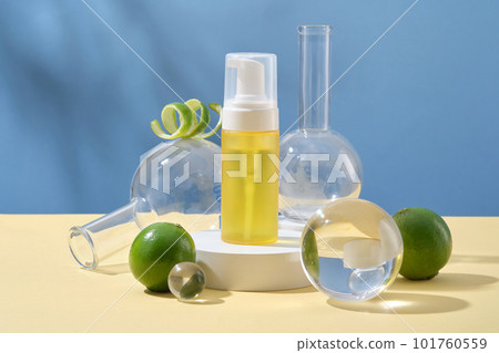 Front view of yellow bottle unbranded on round podium, lab glassware, glass ball and fresh limes decorated on blue background. Cosmetic product mockup extracted from limes 101760559