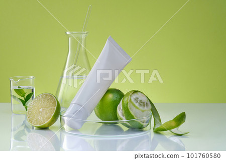 Front view of a white plastic tube unlabeled, fresh limes and lab glassware on green background. Mockup scene for cosmetic, facial cleanser of lime extract with laboratory concept. Space for design 101760580