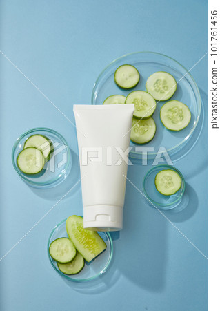An unbranded plastic tube and cucumber slices in petri dish on blue background. Mockup scene for cosmetic of cucumber extract, cleanser or moisturizer, deep cleans and hydrates the skin 101761456