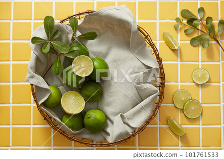 Top view of fresh limes and slices in bamboo basket with gray cloth and green leaves on yellow tile background. Scene for advertising cosmetic with lime ingredient. Empty space to placed product. 101761533
