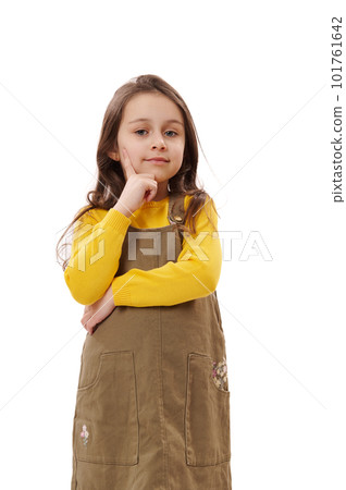 Vertical studio portrait of 5-6 years Caucasian adorable kid girl in khaki and yellow dress, holding finger on her chin and reasoning seriously, looking at camera, isolated on white background. Vertical studio portrait of 5-6 years Caucasian adorable kid girl in khaki and yellow dress, holding finger on her chin and reasoning seriously, looking at camera, isolated on white background. 101761642