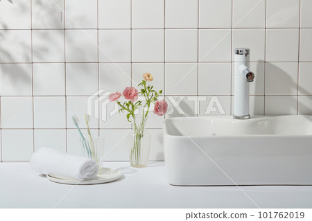 Interior of a modern bathroom with a white tile wall, washstand, towel and toothbrushes. Glass vase with pink flowers that stand out on white background. The concept of bathroom, copy space Interior of a modern bathroom with a white tile wall, washstand, towel and toothbrushes. Glass vase with pink flowers that stand out on white background. The concept of bathroom, copy space 101762019
