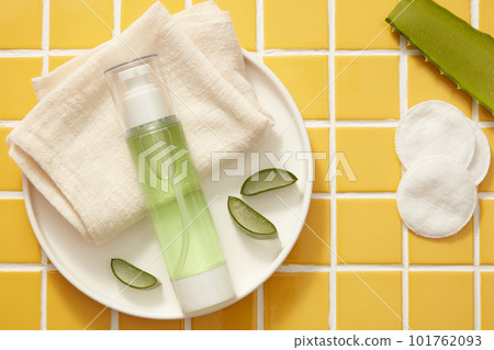 A round dish with folded towel, Aloe vera slices and a pump bottle containing green liquid, decorated with cotton pads. Aloe vera (Aloe barbadensis miller) has many benefits for skin and hair 101762093