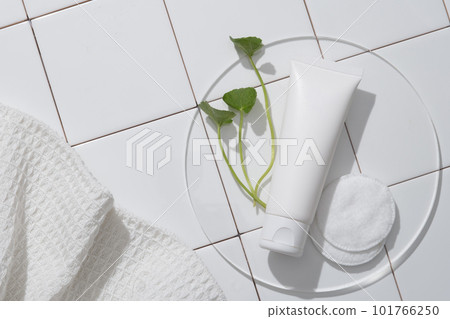 Mockup of a cosmetic product - face cleanser with an unlabeled white plastic tube, gotu kola leaves on a clear podium, and a cotton pad against a background of white tiles. Mockup of a cosmetic product - face cleanser with an unlabeled white plastic tube, gotu kola leaves on a clear podium, and a cotton pad against a background of white tiles. 101766250