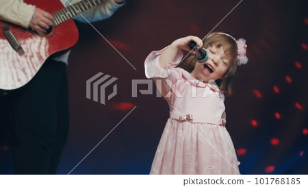 Little girl on stage in vintage dress, she sings into microphone and dances, her father plays acoustic guitar. Color music is shining and smoke is billowing. Father and Child Day, performance on stage Little girl on stage in vintage dress, she sings into microphone and dances, her father plays acoustic guitar. Color music is shining and smoke is billowing. Father and Child Day, performance on stage 101768185